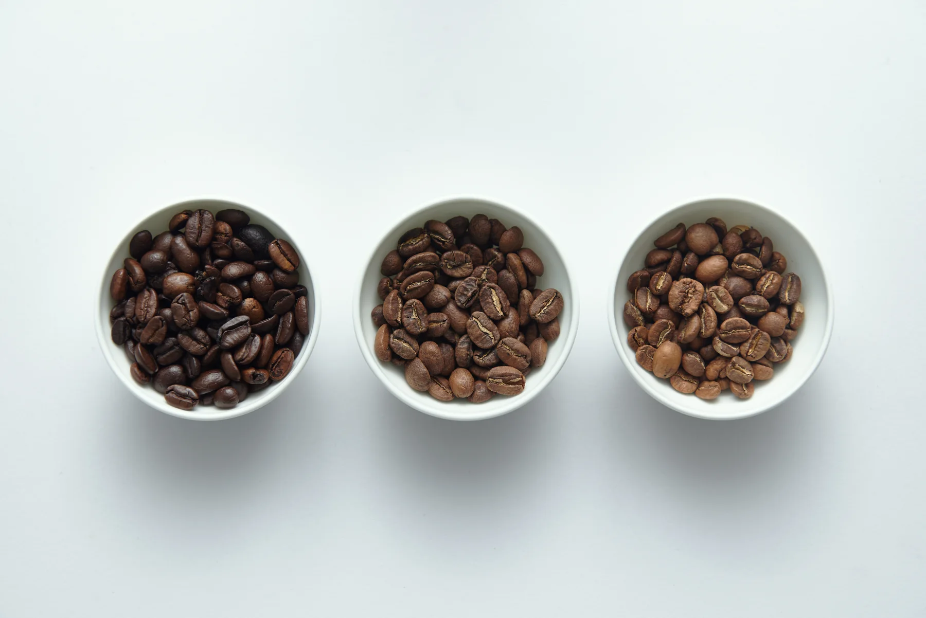 Three bowls containing coffee beans roasted to visibly different degrees on a white surface.