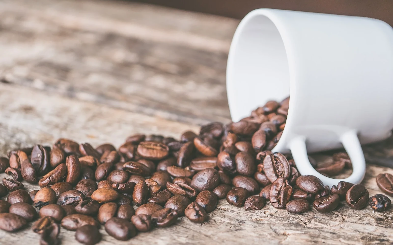 Roasted coffee beans beside a white cup on a wooden table